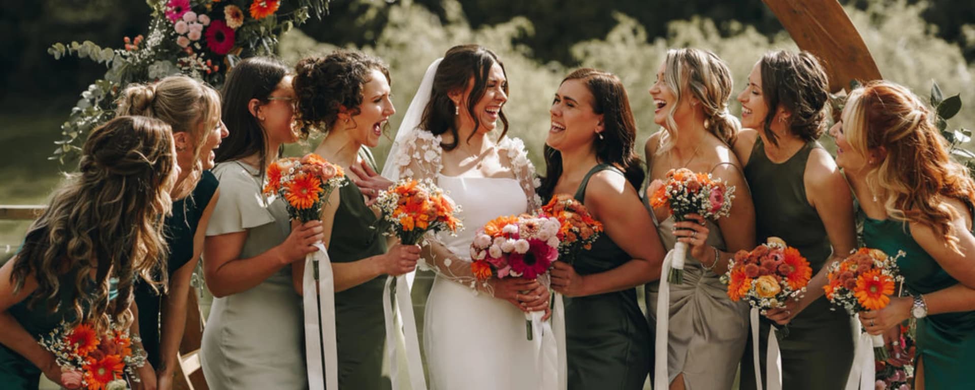 Bride laughing with bridesmaids in green dresses holding bright orange bouquets at an outdoor wedding ceremony.