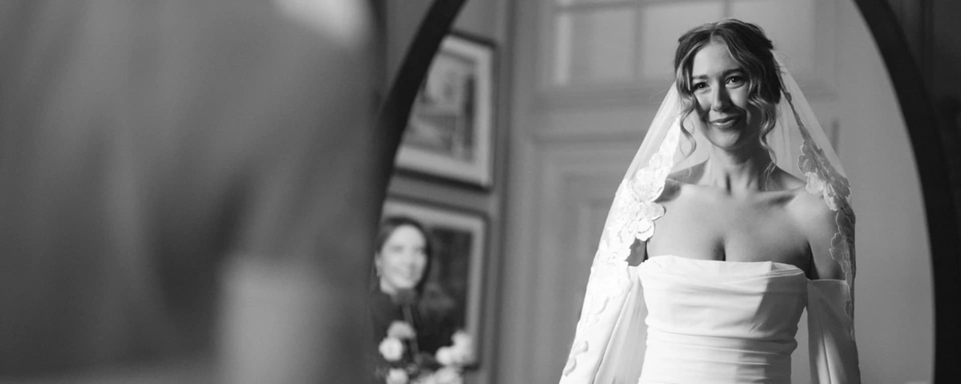 Black and white portrait of bride in veil and soft bridal makeup reflected in mirror before wedding ceremony.