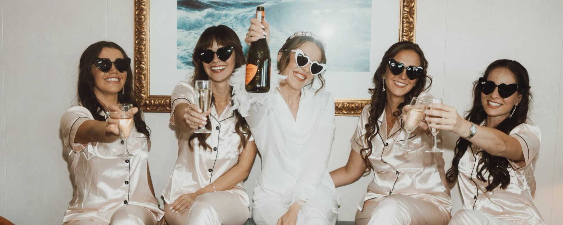 Bride and bridesmaids in satin pyjamas and heart sunglasses raising champagne during wedding morning preparations in London.